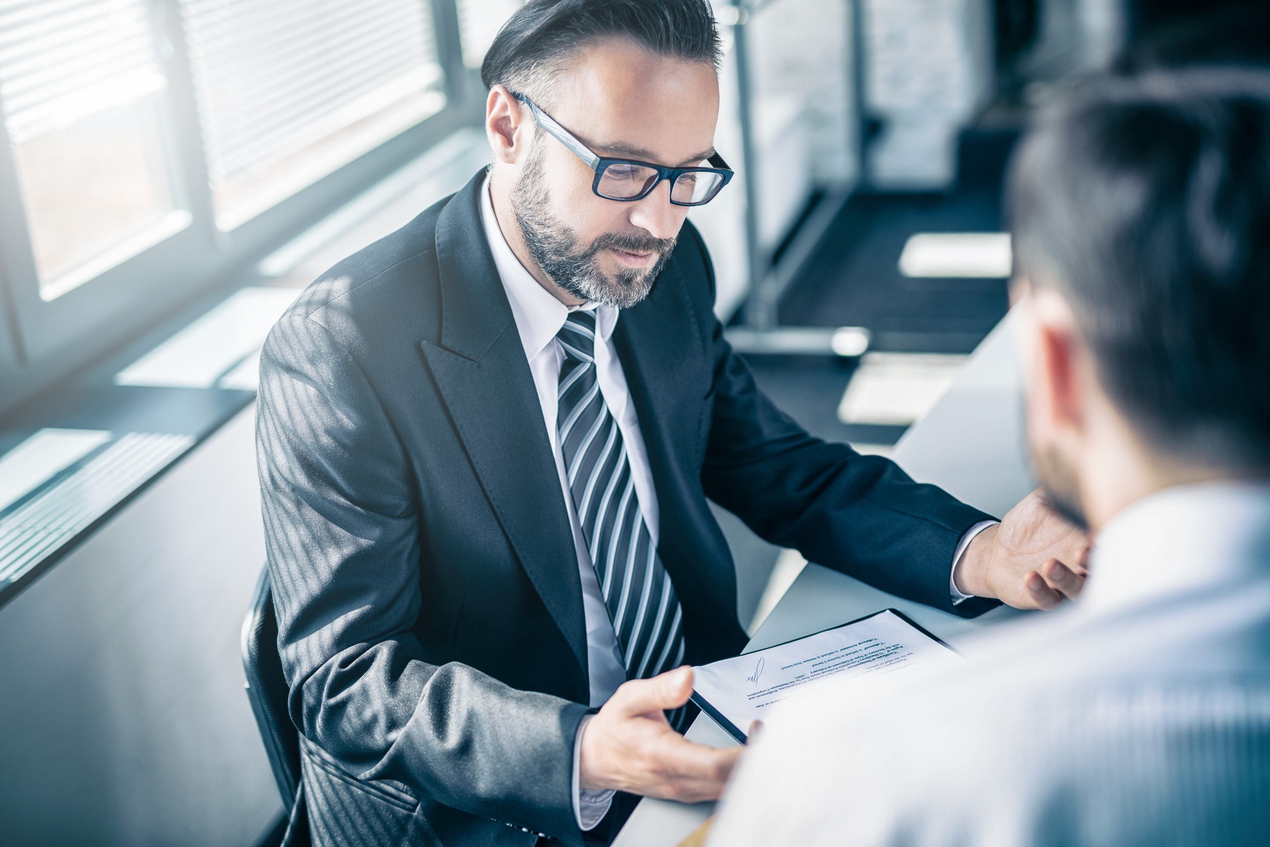 A man in a suit and glasses, likely an independent insurance broker, discusses a contract with another person across a desk in a bright office setting.