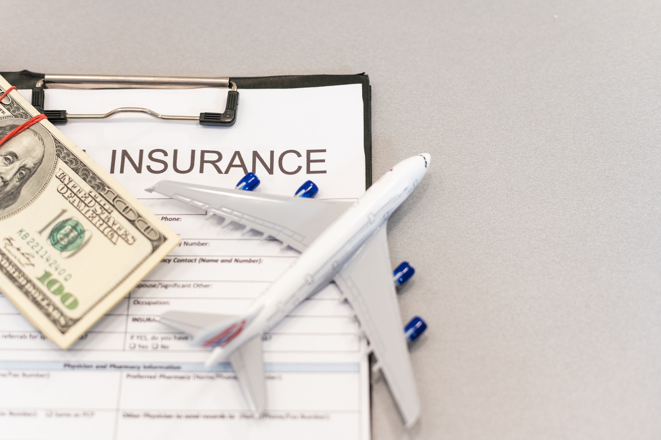 A clipboard with an insurance form, a stack of US dollar bills, and a model airplane placed on top, representing Independent Insurance Coverage and travel expenses for Minnesota travelers.